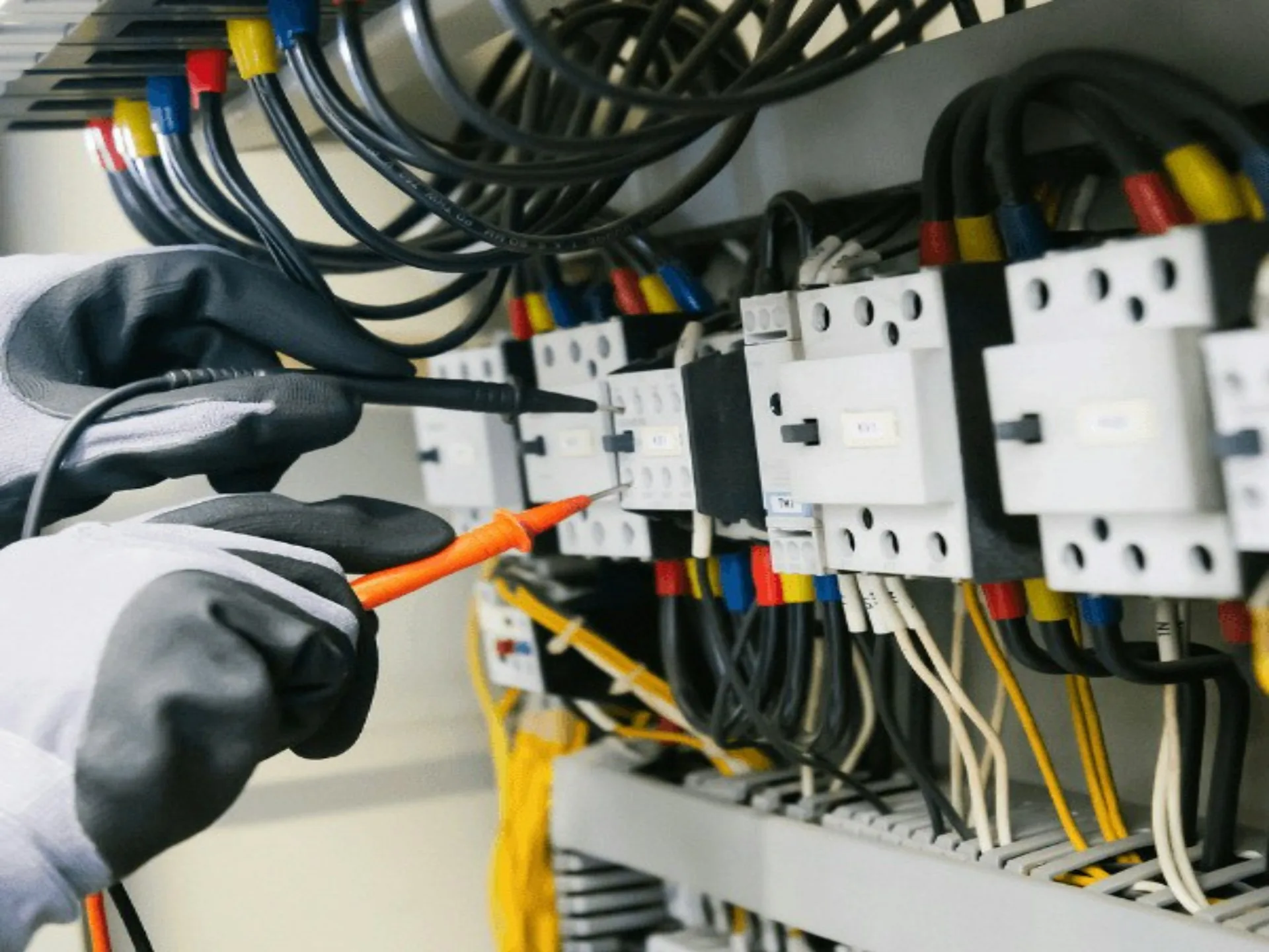 Gloved hands testing wiring in an open electrical panel with a multimeter probe.