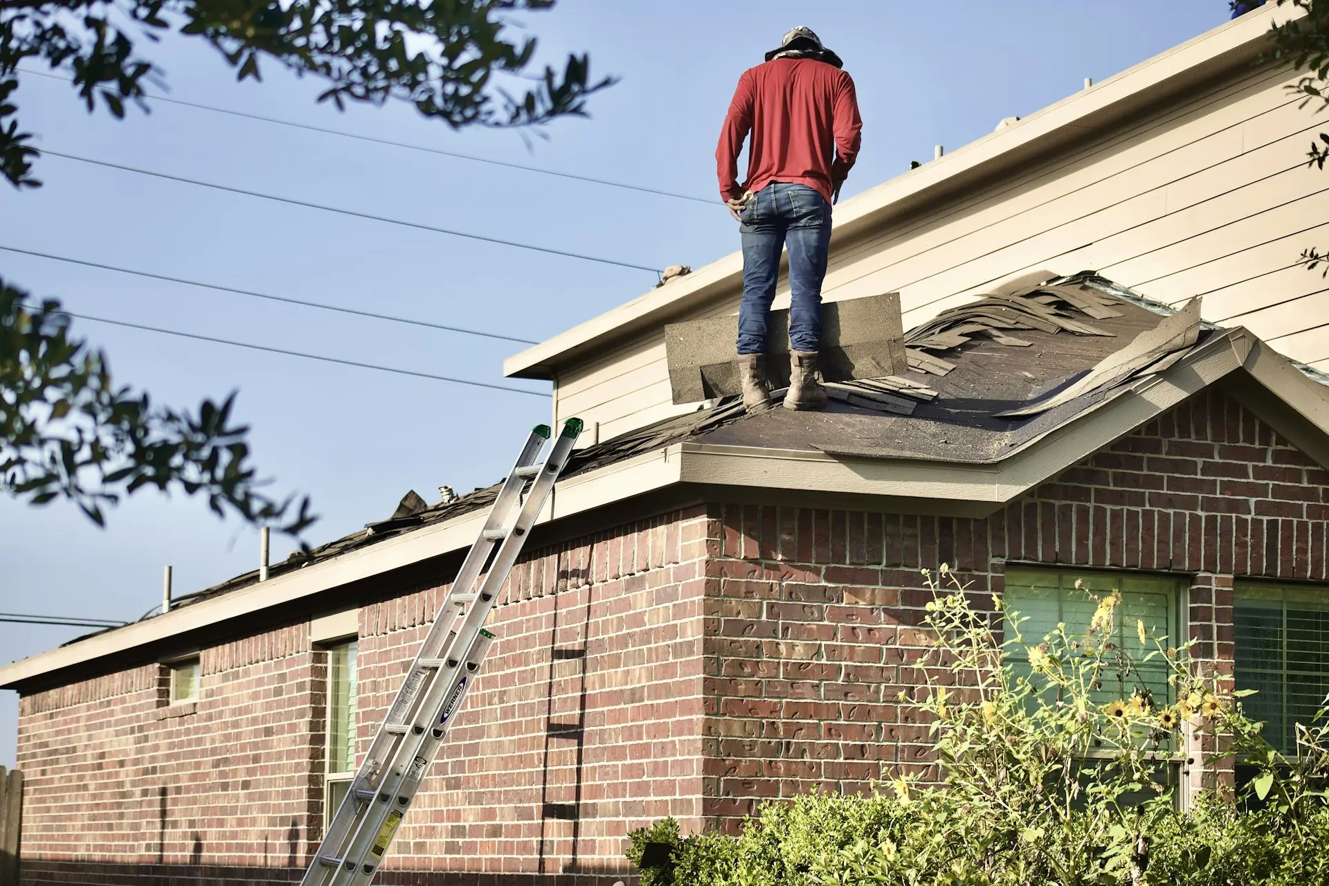 A roofer tearing off old shingles on a single-story brick house, ladder against the eave.